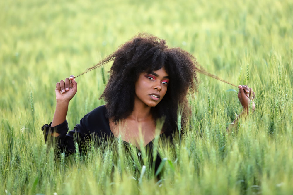 Woman with long natural hair sitting calmly in a grassy field, holding and gently pulling her hair with both hands.