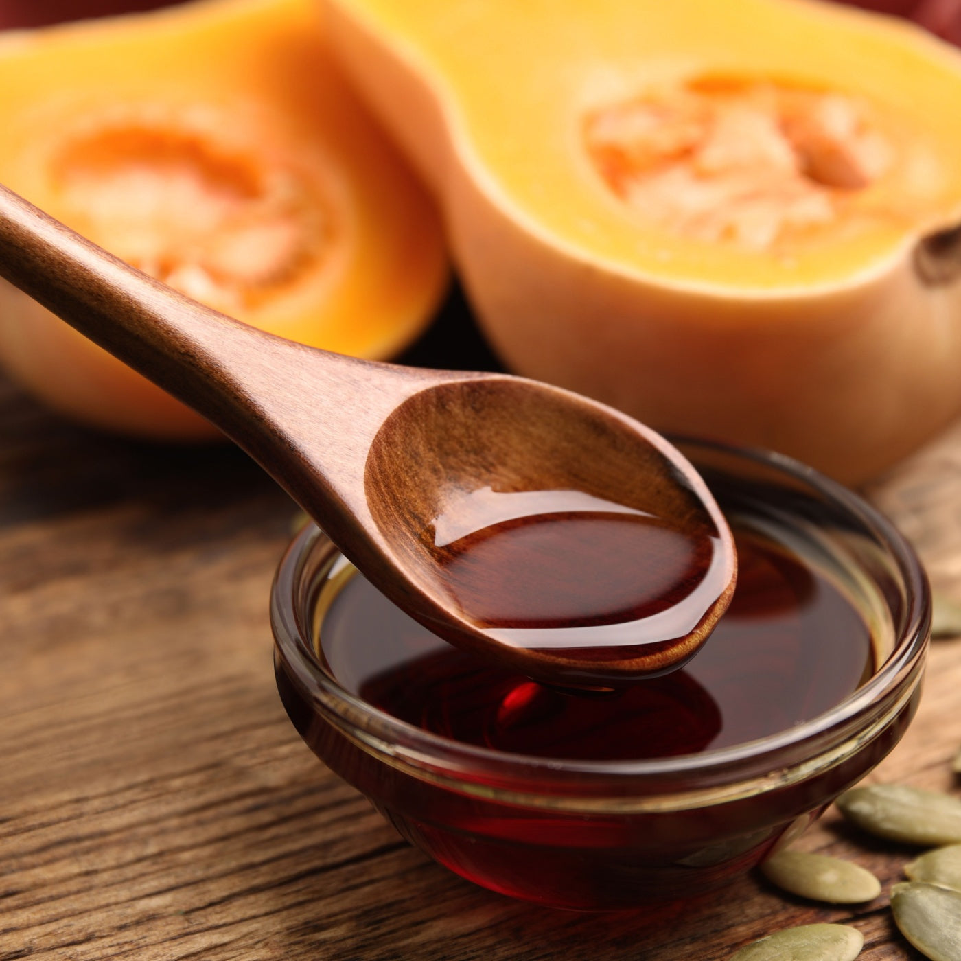 Spoon with pumpkin oil over bowl and seeds on wooden table, closeup